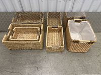 Four wicker baskets placed on concrete floor against metal wall. Two nested rectangular baskets with metal handles, one small rectangular basket, one taller rectangular basket with white fabric lining and wooden handles.