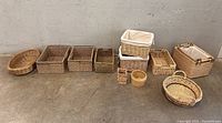 Photo of assorted wicker and rattan baskets arranged in a row on a concrete floor, against a light-colored wall.