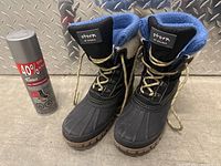 Front view of pair of black Storm brand winter boots with blue knit cuff and beige sherpa lining, placed next to a can of 400% Tanax water protector spray.