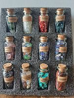 Front view of a set of 12 small glass bottles filled with different gemstone chips, each labeled with its stone name, arranged in a black foam insert.