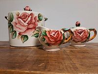 Front view of hand-painted ceramic canister, creamer, and sugar bowl showing rose artwork and lid details