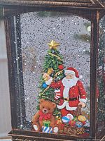 Close-up of lantern scene showing Santa Claus, Christmas tree decorated with gold star and garlands, teddy bear, and gifts inside the lantern with glitter background.