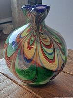 Front view of large art glass vase showing colorful swirled pattern and ruffled blue rim, placed on wooden surface near window with natural light.