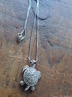 Silver-colored turtle pendant necklace with detailed textured shell and moveable head and limbs on a 16-inch chain lying on a wooden surface.