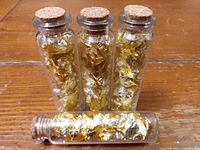 Four small glass vials filled with gold leaf flakes with cork stoppers on a wooden surface.
