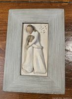 Top view of the memory box lid showing the carved mother and daughter figurine relief in soft clays or resin shades, surrounded by a textured gray wooden frame.