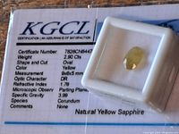 Natural yellow sapphire loose stone in a protective display box with KGCL certificate underneath detailing stone's specifications such as weight (2.90 Cts), shape (Oval), color (Yellow), measurements (9x6x5 mm), refractive index (1.76) and species (Corundum).