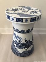 Front view of the porcelain garden stool showing the blue and white painted horsemen and mountain scene on the body and patterned band around the edges.