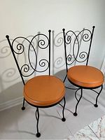 Two vintage bistro chairs with black iron frames and round orange vinyl seats placed side by side against a white wall.