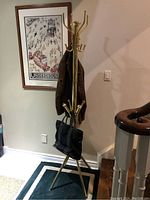 Full view of a tall brass coat tree holding a coat and a black handbag, positioned by a framed wall poster and staircase.