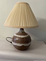 Angled front view of the complete table lamp with pleated beige fabric lampshade attached, showing ceramic base with brown and white glaze.