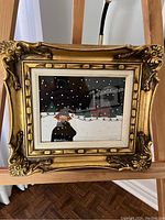 Front view of vintage gold ornate frame containing mixed media fabric art portraying a winter scene with a figure and house amidst falling snow.