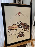 Full view of the framed First Nations painting featuring a moose and birds on an easel.