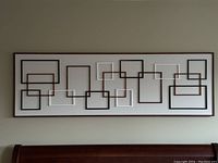 Front view of the geometric squares wall art showing overlapping black, white, and brown squares on a white background in a thin dark frame above a wooden headboard.