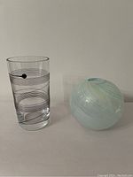 Two Kosta Boda vases shown side by side, clear tall one with black linear/dot design, and round swirled blue globe vase.