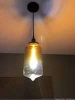 Lit clear glass pendant ceiling light fixture mounted on ceiling, casting warm light with visible filament bulb inside, showing adjustable cord and black mount.