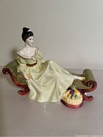 Side view of Royal Doulton porcelain figurine showing woman seated on green and red chaise lounge with fruit basket at footrest.