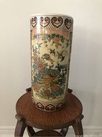 Full view of cylindrical cream ceramic umbrella stand with intricate painted Asian motifs placed on wooden stool.