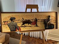 Full view of rectangular mid century wall art with wooden frame, fabric background depicting bullfighting arena and ceramic figures including a matador, charging bull, and picador on horse.