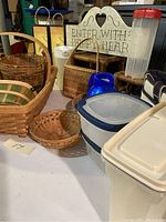 Photo showing assortment of woven baskets including picnic basket, shallow basket, woven rectangular tray, and containers including blue ceramic pitcher, plastic pitcher with red lid, and plastic food storage containers.