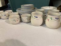 View of multiple stacked dinner plates, cups, bowls, and two larger serving vessels featuring a floral vine pattern in muted colors on white ceramic.