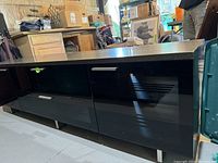 Front view of large black entertainment unit showing smooth top, two black-tinted glass doors with silver handles, and metal legs.