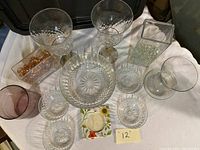 Photo showing twelve glassware items including goblets, bowls, a purple tinted container, and decorative glass pieces on white cloth.