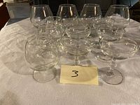 Photo showing an assortment of larger brandy snifters, champagne coupe glasses, and cordials arranged on a cloth surface.