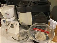 Photo showing white Hamilton Beach drip coffee maker, hand mixer, popcorn maker, clear glass dishes, and wooden paper towel holder in garage setting.