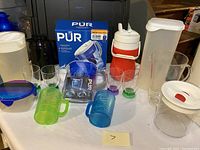 Photo showing an assortment of kitchen plastic items including PUR water filter in box, colorful plastic cups, thermos, and pitchers.