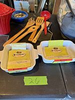 Two Le Creuset ceramic baking dishes with labels, wooden kitchen utensils, red ceramic teapot or sugar bowl with lid, metal strainer with blue rim on a black surface