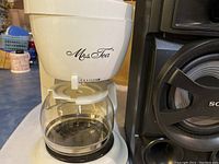 White Mrs. Tea coffee maker with clear glass carafe sitting on a blue countertop.