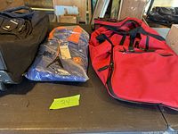 Set of travel bags on table including black rolling suitcase, red duffel bag, and blue packaged item.