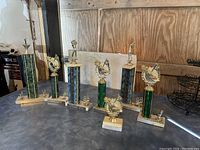 Photo showing six golf trophies with gold-tone golfer figure toppers and golf motifs on green and blue columns, mounted on rectangular marble bases in a garage setting.