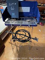 Blue two burner camp stove on foldable metal stand with coiled hose underneath, showing the stove setup from front angle.