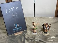 View of two Hummel figurines placed on a cloth surface with a lot number in the background