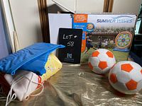 Photo showing the Summer Waves inflatable soccer target packaging box, two soft soccer balls with orange hexagon design, and a blue and yellow air pump inflator.
