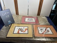 Photo of three framed Jackson Beardy prints displayed on a table with different colored mattes: pink, mustard yellow, and orange.