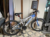 Side-angle photo of white and blue Nakamura Solano 6.2 mountain bike showing entire bike, tires, frame, seat, pedals, and handlebars on a tiled floor indoors.