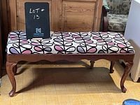 Wooden bench with upholstered seat covered in white fabric with black and pink leaf pattern, viewed from front against wooden background.