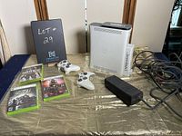 Wide shot showing Xbox 360 console, two controllers, three game cases, power adapter, and cables against tan textured surface