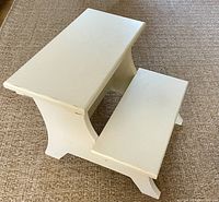 Image showing top view of antiqued-white two-step wooden stool on woven rug, highlighting size and flat step surfaces.