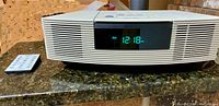 Front view of white Bose Wave Radio/CD Player with digital clock display showing time 12:18 PM and FM radio indicator, placed on dark granite surface, with remote control beside it.