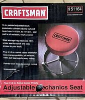Box front showing product image and detailed specifications for Craftsman Adjustable Mechanics Seat including padded 12-inch seat, pneumatic cylinder for height adjustment, steel storage tray, 5 swivel caster wheels and powder coat finish.