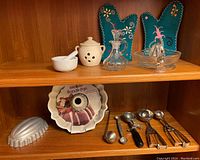 Shelf with vintage ceramic and glass kitchenware including garlic pot, mortar and pestle, oil and vinegar cruet, lemon juicer, Bundt pan, jello mold, ice cream dishers, melon baller, ladle, and embroidered oven mitts.