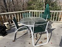 Photo of a 4 ft diameter round glass patio table with white metal frame, surrounded by 4 green sling fabric chairs with white metal frames, and a closed green patio umbrella on a wooden deck outdoors.