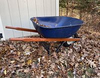 Blue steel wheelbarrow with brown hardwood handles on the ground outdoors, showing overall structure and condition.