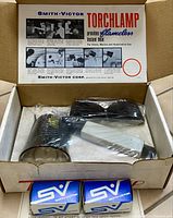 Open box showing the Smith-Victor Torchlamp with torch head and white handle, two blue and white branded accessory boxes, and a pair of protective glasses wrapped in plastic. The inside lid of the box has printed instructions.