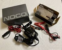 NOCO Genius2 trickle charger with clamps, power cord, and original box on a fabric surface.