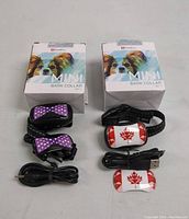Two Mini Bark Collars with USB charging cables and boxes. One collar black with purple bow tie design, the other white with red maple leaf design.